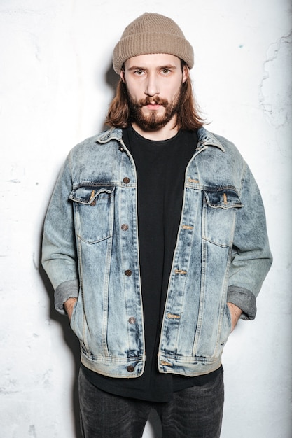 attractive bearded hipster man wearing hat dressed in jeans jacket posing over wall