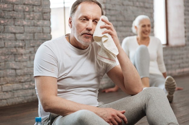 Attractive bearded man keeping towel near his eye putting arms on the legs while sitting on the floor