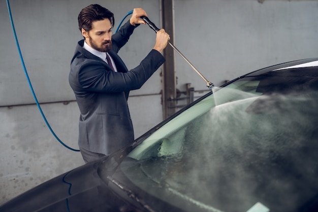 Uomo laborioso concentrato alla moda attraente barbuto in vestito che pulisce la sua automobile alla stazione di self service di lavaggio manuale dell'automobile.