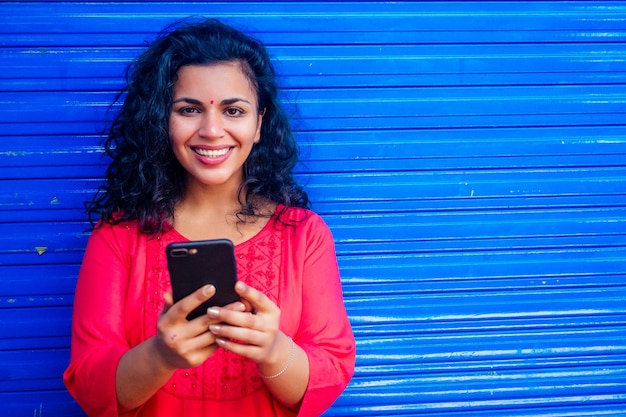 Attractive beautiful happy young latin hispanic woman with teak bindi on forehead smiling on blue wall street background holding smartphone modern