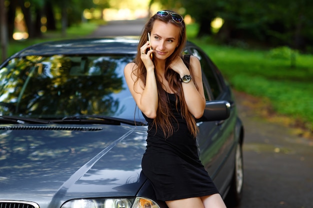 Attractive beautiful woman in black dress talking on smart phone near the car