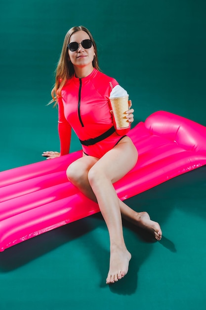 Attractive beautiful woman slim body in a bright pink swimsuit in sunglasses lies on an inflatable pink mattress isolated on a green studio background Summer vacation at the sea tanning concept