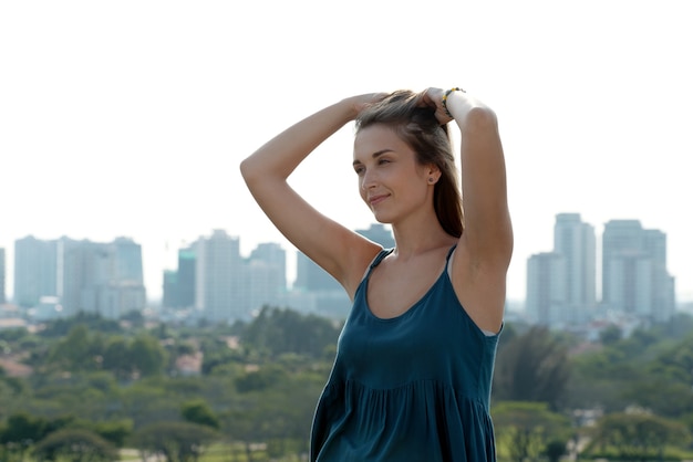 Attractive beautiful young woman touching her hair and looking at big city when standing on building...