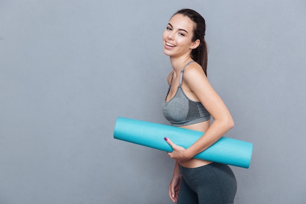 Attractive beautifull young sportswoman holding yoga mat isolated over grey background