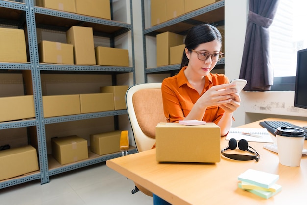 Foto attraente bellezza ragazza proprietario dello shopping online utilizzando lo smart phone mobile che controlla l'ordine del cliente e seduto sulla scrivania del magazzino preparando la scatola di spedizione.