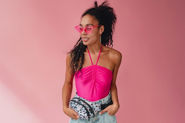 Attractive black african american woman in stylish summer outfit pink top and shorts bum bag on pink studio background summer fashion trend happy smiling curly hair sunglasses accessories