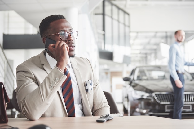 Attractive black businessman sits at the table at the car dealership, he signs a contract and buys a new car.