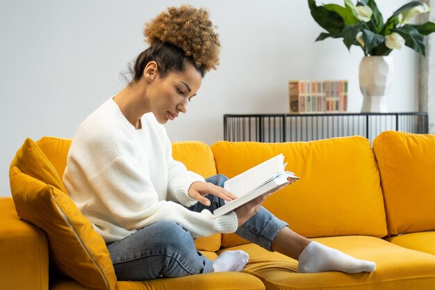 Attractive black curlyhaired woman reading her favorite historical novel sitting on a cozy yellow