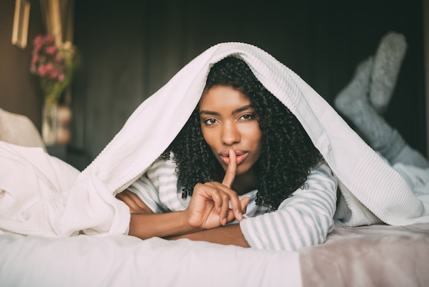 Attractive black woman asking for silence with finger on lips on bed