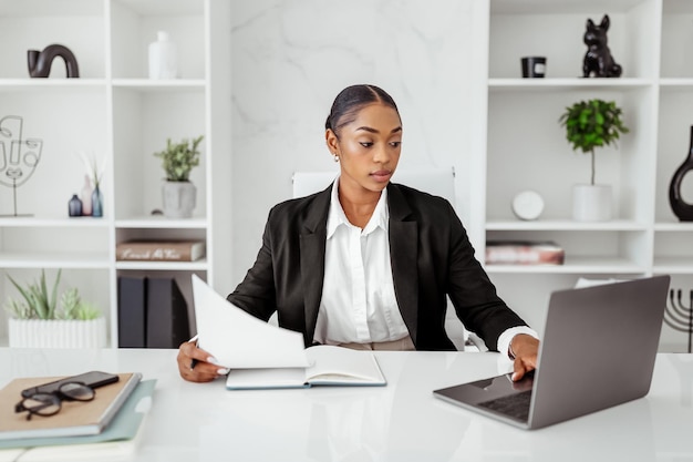 Attractive black woman manager in formal wear sitting at table with laptop and checking papers