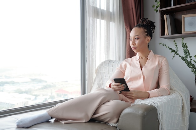 Attractive black young woman sitting in big chair buy window holding smartphone and looking away