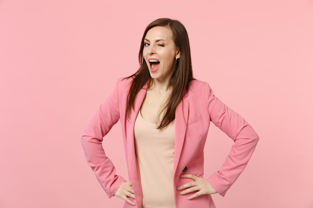 Attractive blinking young woman in jacket keeping mouth wide open, standing with arms akimbo on waist isolated on pastel pink background. People sincere emotions lifestyle concept. Mock up copy space.