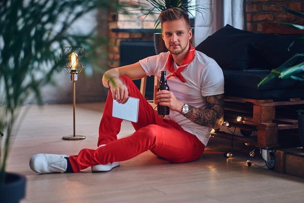 Attractive blond male with tattoos on arms, dressed in a red Christmas pants using tablet PC in a room with loft interior.