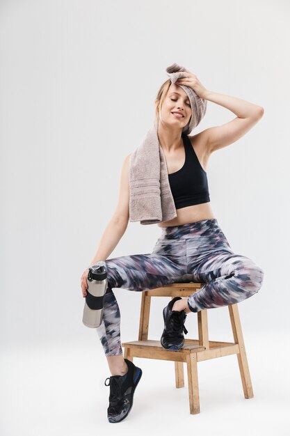 attractive blond woman  dressed in sportswear with towel over neck sitting on chair after workout in gym isolated over white wall