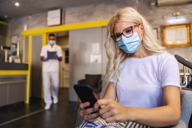 Photo attractive blond woman with face mask sitting in hospital, using mobile phone and waiting to be called by a doctor.