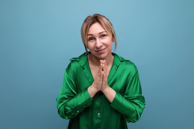Attractive blond young woman in green silk shirt praying on blue background with copy space