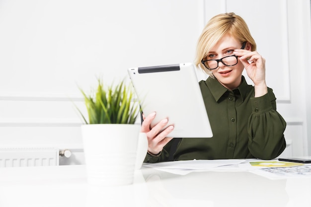 Attractive blonde businesswoman wears green shirt and eyeglasses work with tablet computer in white office room