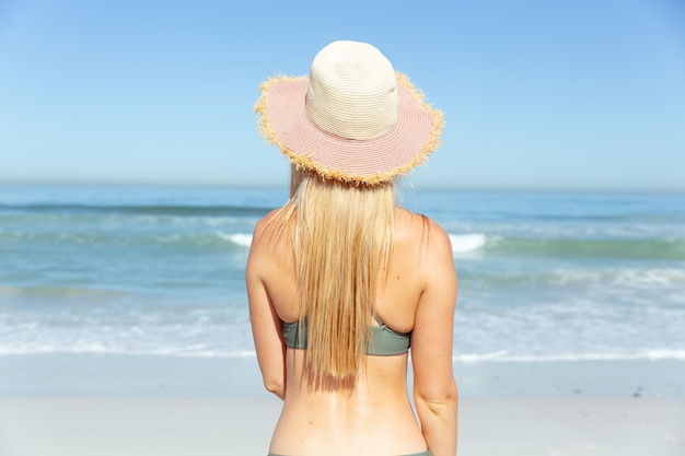 Attractive blonde caucasian woman enjoying time at the beach on a sunny day, wearing a sun hat, facing sea, with blue sky and sea in the background. summer tropical beach vacation