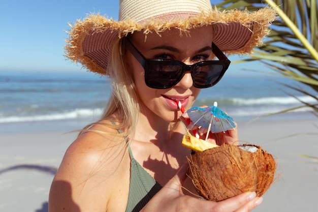 Привлекательная кавказская блондинка, наслаждающаяся временем на пляже в солнечный день, в шляпе от солнца, держащая коктейль и пьющая, с голубым небом и морем на заднем плане. Летний тропический пляжный отдых.