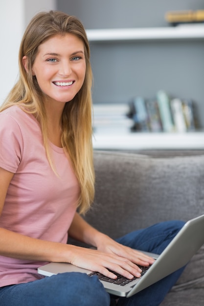 Attractive blonde sitting on her sofa using laptop looking at camera