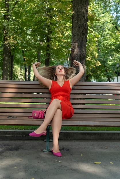 공원에서 벤치에 매력적인 금발의 여자 빨간 드레스에 젊은 여자 여름 날 공원에서 이완 세로 프레임