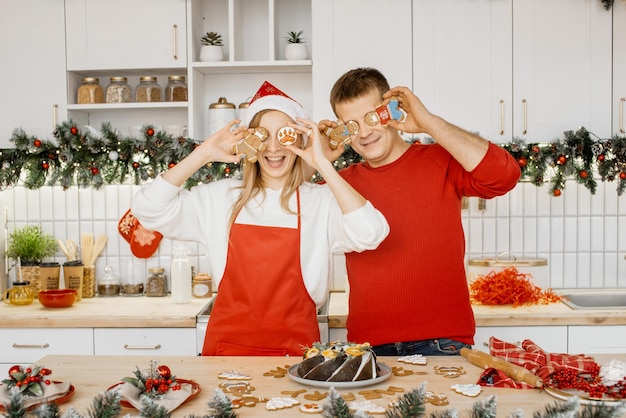 写真 キッチンで赤いニットのセーターを着た夫の隣に、サンタの帽子と赤いエプロンを着た魅力的な金髪の女性が、手作りのジンジャーブレッド・クッキーを楽しんでいます