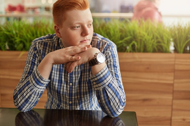 An attractive boy with red hair dressed in checked elegant shirt sitting in cosy cafe with green plantation holding hands
