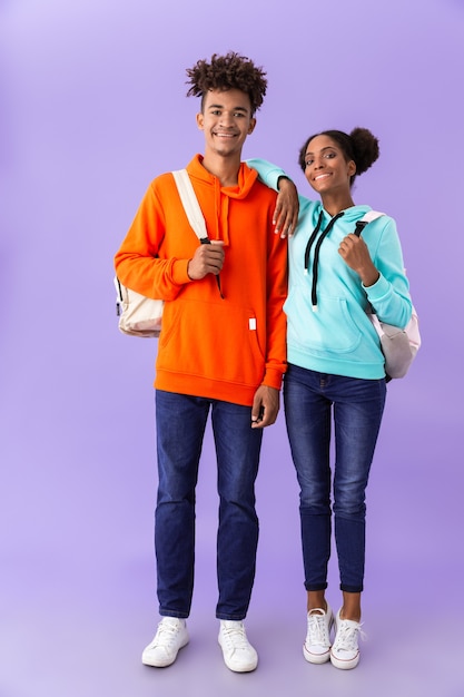 Attractive brother and sister wearing backpacks hugging together, isolated over violet wall