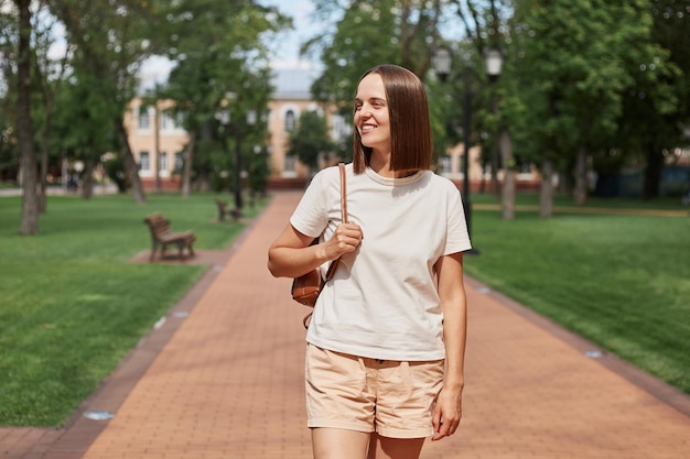 Attractive brown haired girl resting strolling pastime on fresh air outdoors wearing white tshirt and holding backpack walking on path in city park