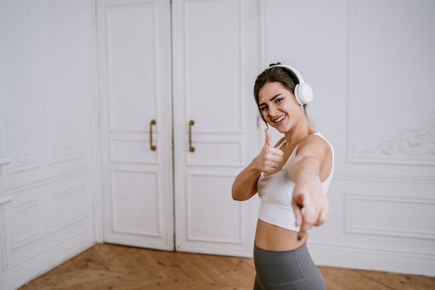 Attractive brunette American girl in headphones sportswear points at camera shows thumb up gesture