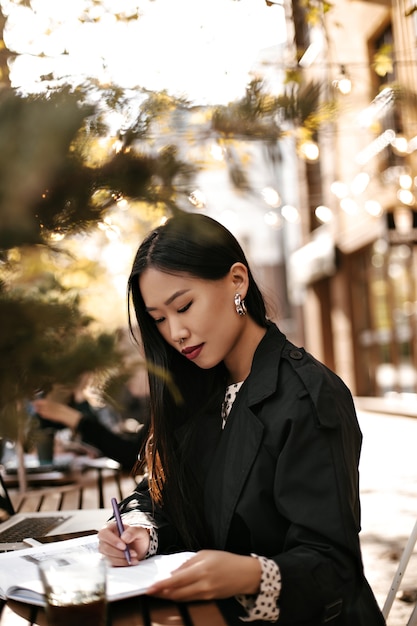 Attractive brunette Asian woman in stylish black trench coat sits outside, makes notes in her notebook