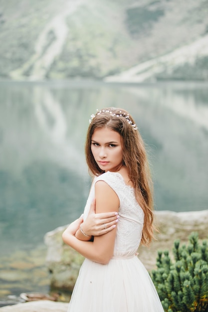 Foto attraente sposa bruna in splendido abito da sposa bianco in piedi vicino al lago morskie oko