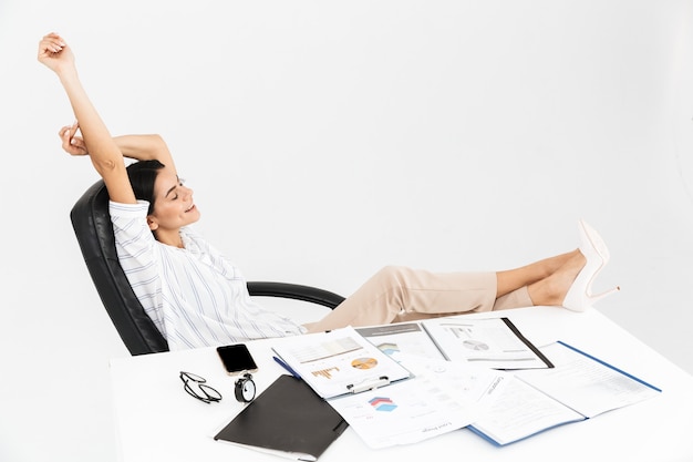 attractive brunette businesswoman smiling while sitting in armchair at table and working in office isolated over white wall