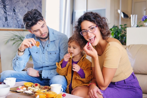 Attractive brunette female in eyeglasses, bearded stylish male and their cute little daughter sits on a couch and eat lunch in a living room.