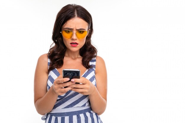 attractive brunette girl in a striped dress and glasses with a phone in her hands on a white wall