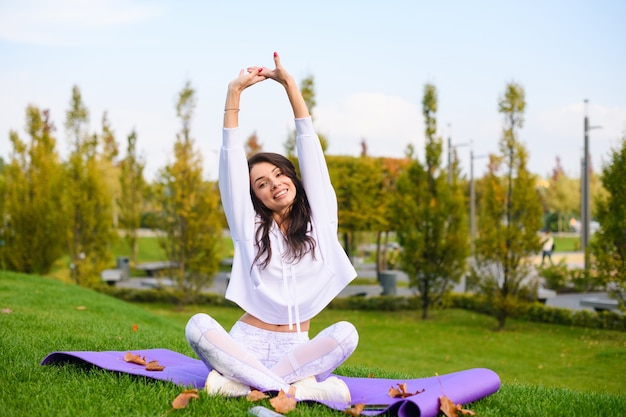 Attractive brunette girl in white sportwear sit on purple mat in lotus pose and stretch up hands under head, outdoor fitness, yoga exercises