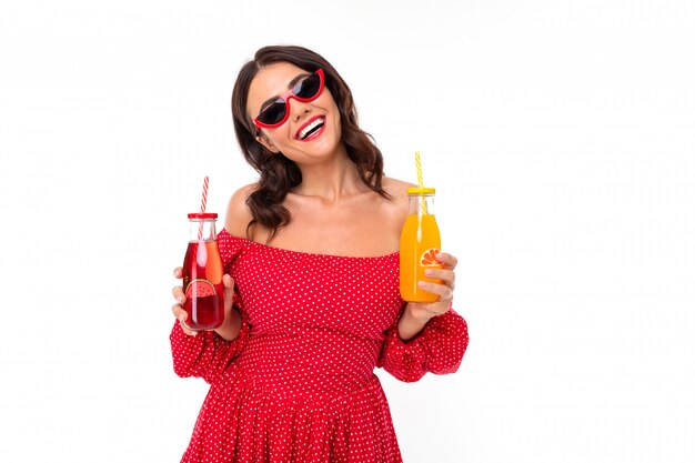 привлекательная брюнетка с очками в красном платье с бокалами коктейля на белой стене