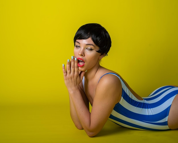 Attractive brunette lies on her stomach in a striped bikini shorthaired young woman in bodysuit on a yellow background in the studio