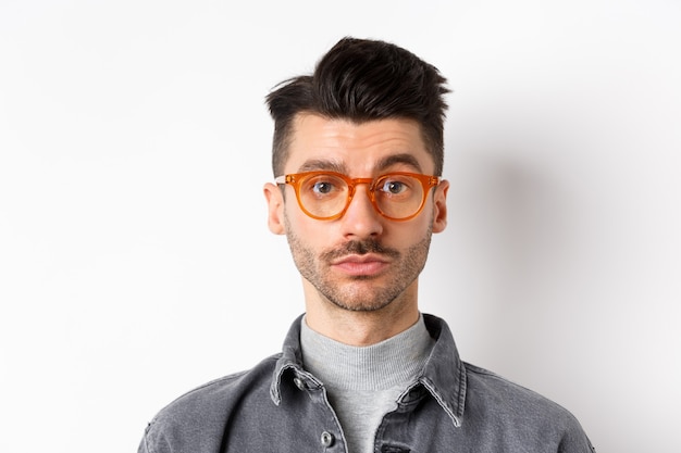 Attractive brunette man with moustache, wearing stylish glasses and look serious at camera, no emotion pokerface, standing against white background.