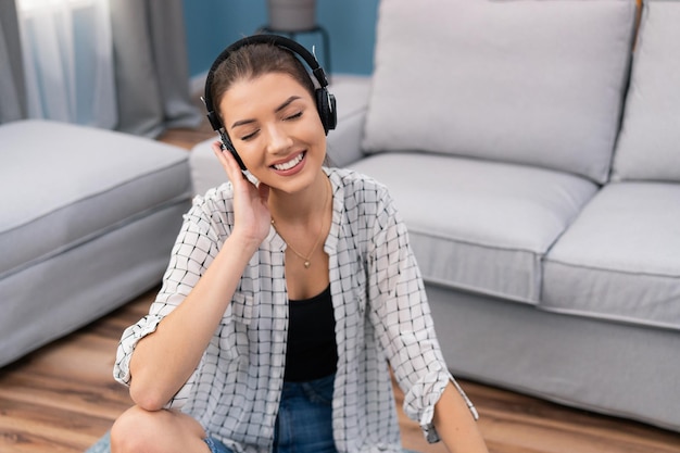 An attractive brunette sits on the living room floor with wireless headphones on ears