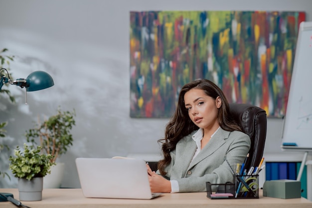 An attractive brunette with dark eyes wearing jacket sits in a comfortable chair in front of laptop