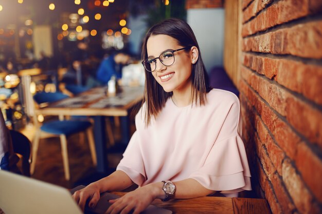 Attractive brunette with eyeglasses using laptop while sitting in cafeteria