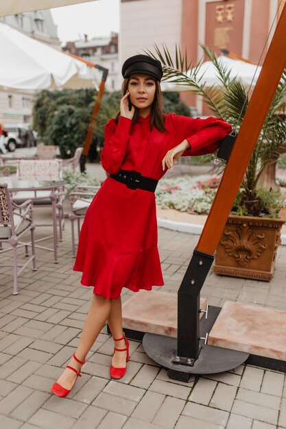 Attractive brunette with short hair with gaze standing on street Fulllength photo of girl in casual autumn bright dress and red shoes