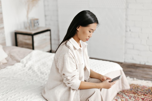 Attractive brunette woman in beige cardigan and pants siting on soft bed and messaging in phone Portrait of Asian lady in stylish outfit poses in bedroom