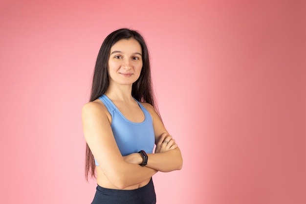Attractive brunette woman in fashionable sportswear on pink background Healthy lifestyle concept Happy face smiling with crossed arms looking at the camera Positive person