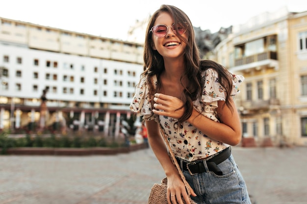 Attractive brunette woman in jeans trendy floral blouse and pink sunglasses smiles sincerely walks outdoors and looks into camera