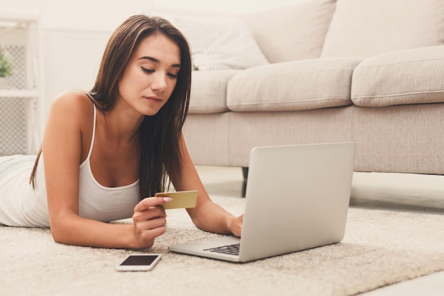 床のカーペットの上に横たわって、自宅でクレジットカードとラップトップでオンラインショッピングをしている魅力的なブルネットの女性。オンラインショッピング、テクノロジー、e-moneyのコンセプト