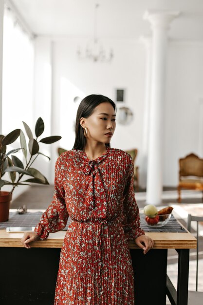 Attraente donna bruna in abito floreale rosso si appoggia sul tavolo di legno bella ragazza asiatica in abito elegante pone in cucina