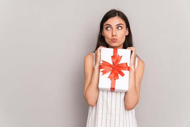  attractive brunette woman wearing dress thinking while holding present box isolated over gray wall