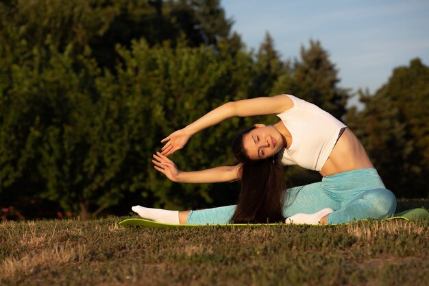 Attractive brunette woman wearing sport apparel practicing yoga in the park at sunset. Space for text